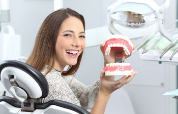 A smiling woman in a dentist chair presenting teeth and gums model