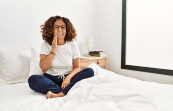 Middle-aged Hispanic woman with a jaw pain caused by night bruxism sitting on her bed after waking up.