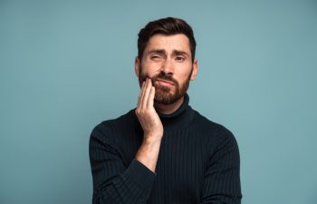 Dental problems. Portrait of unhealthy man pressing sore cheek, suffering acute toothache, periodontal disease, cavities or jaw pain. Indoor studio shot isolated on blue background